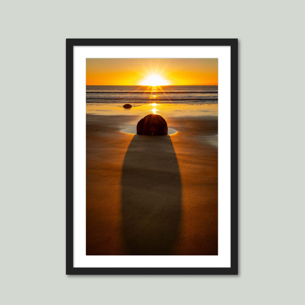 The Patient Stone, a Moeraki Boulder casting a long shadow toward the viewer as the sun rises over the Pacific in Otago, New Zealand, in a black frame with a white mat