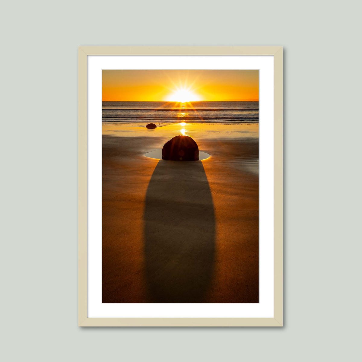 The Patient Stone, a Moeraki Boulder casting a long shadow toward the viewer as the sun rises over the Pacific in Otago, New Zealand, in a maple frame with a white mat