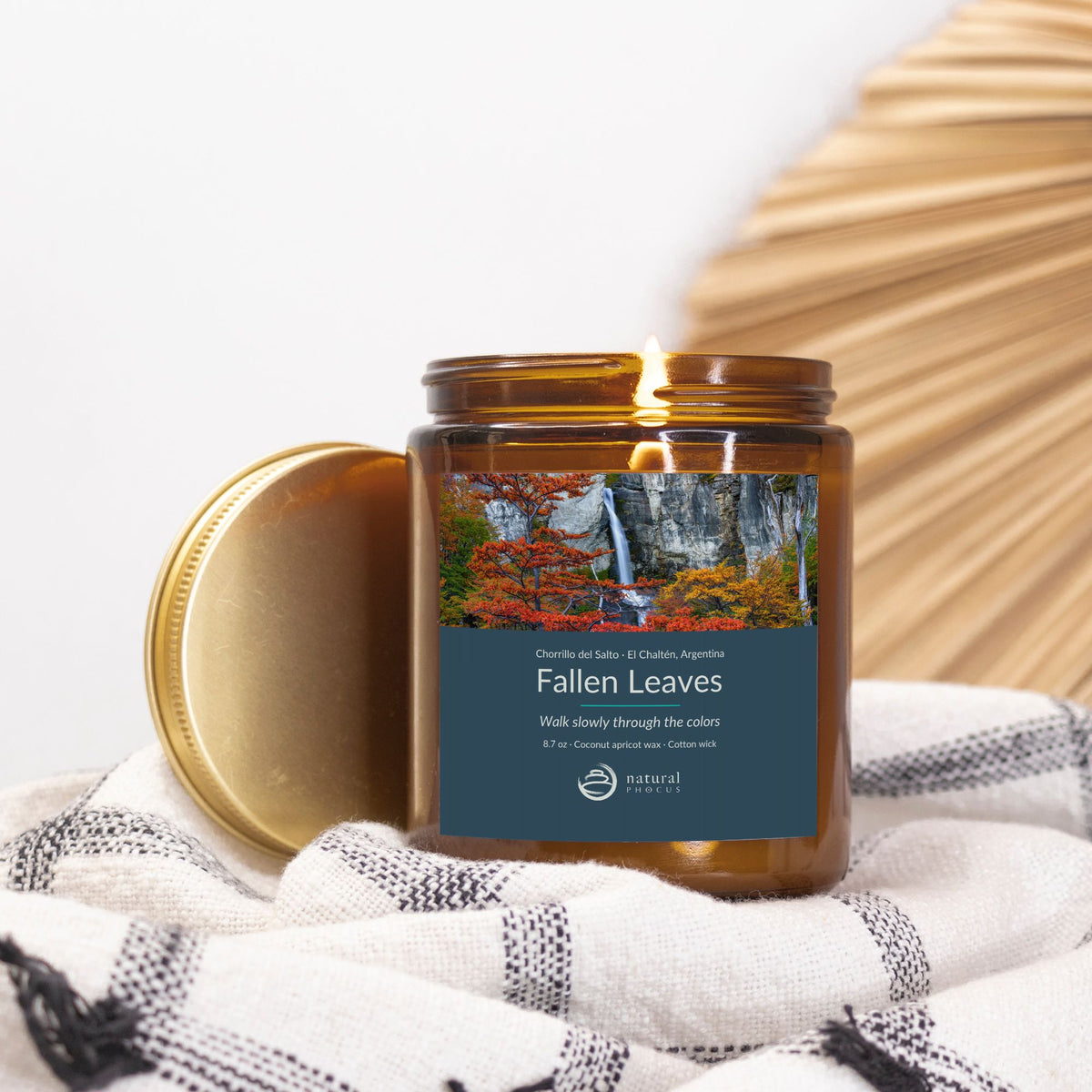 Chorrillo del Salto Candle in Fallen Leaves, an amber glass jar candle with a gold lid, labeled with a photograph of a waterfall framed by red and gold autumn trees near El Chaltén, Argentina