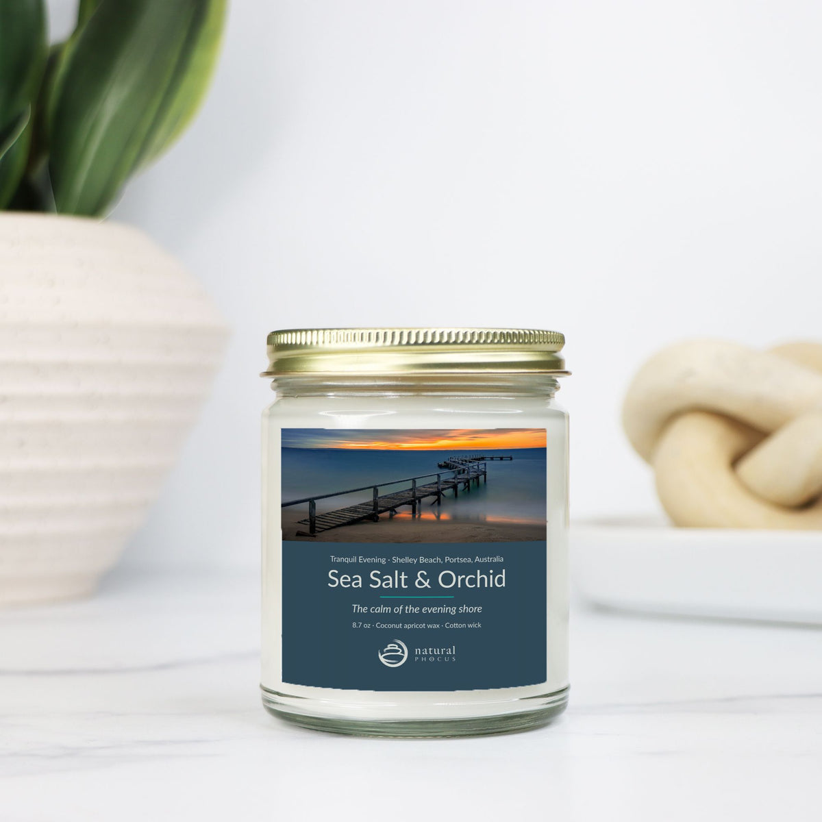Tranquil Evening Candle in Sea Salt and Orchid, a clear glass jar candle with a gold lid, labeled with a photograph of a wooden jetty curving into still water at sunset at Shelley Beach, Portsea, Australia