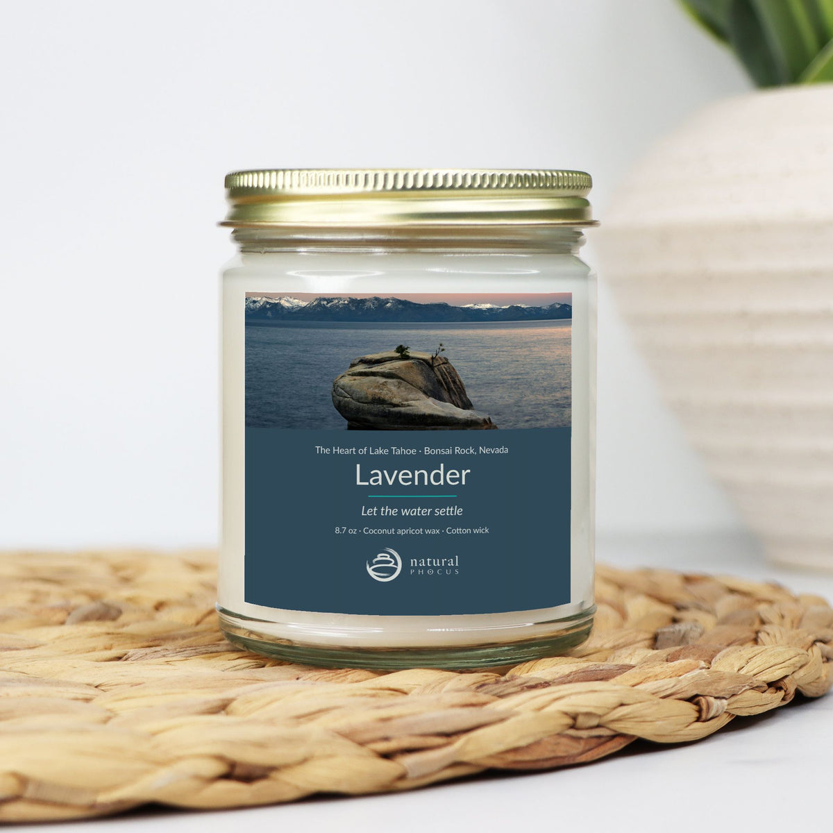 The Heart of Lake Tahoe Candle in Lavender, a clear glass jar candle with a gold lid, labeled with a photograph of Bonsai Rock in still water beneath snow-covered peaks at Lake Tahoe, Nevada