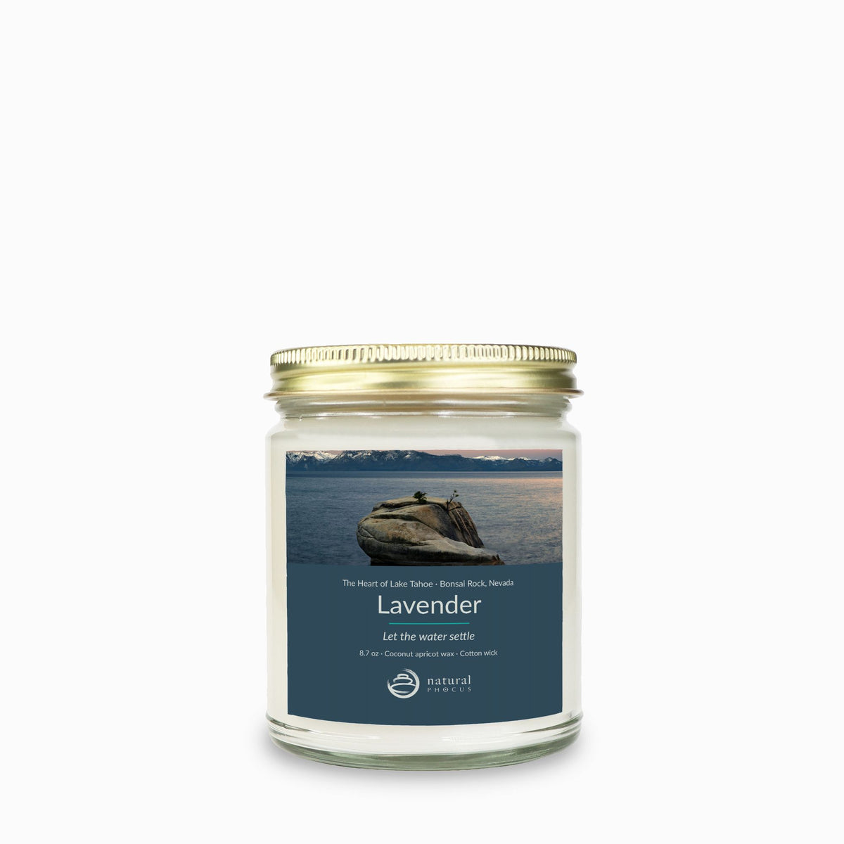 The Heart of Lake Tahoe Candle in Lavender, a clear glass jar candle with a gold lid, labeled with a photograph of Bonsai Rock in still water beneath snow-covered peaks at Lake Tahoe, Nevada