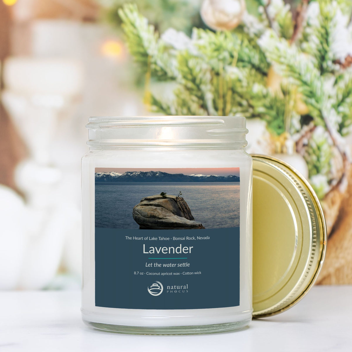 The Heart of Lake Tahoe Candle in Lavender, a clear glass jar candle with a gold lid, labeled with a photograph of Bonsai Rock in still water beneath snow-covered peaks at Lake Tahoe, Nevada