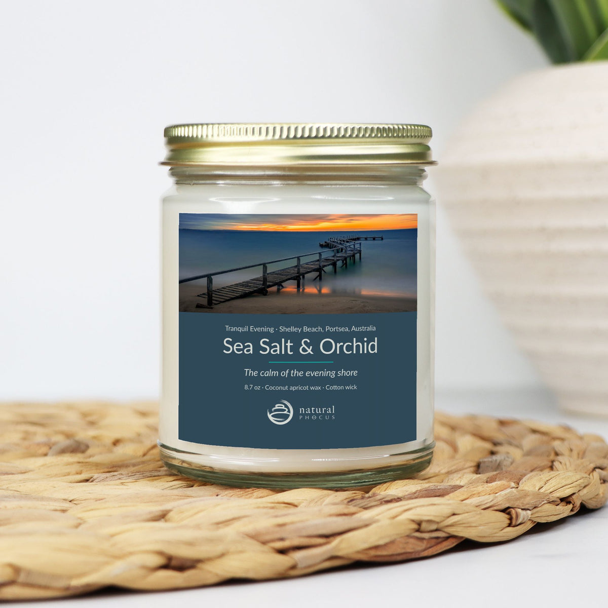 Tranquil Evening Candle in Sea Salt and Orchid, a clear glass jar candle with a gold lid, labeled with a photograph of a wooden jetty curving into still water at sunset at Shelley Beach, Portsea, Australia