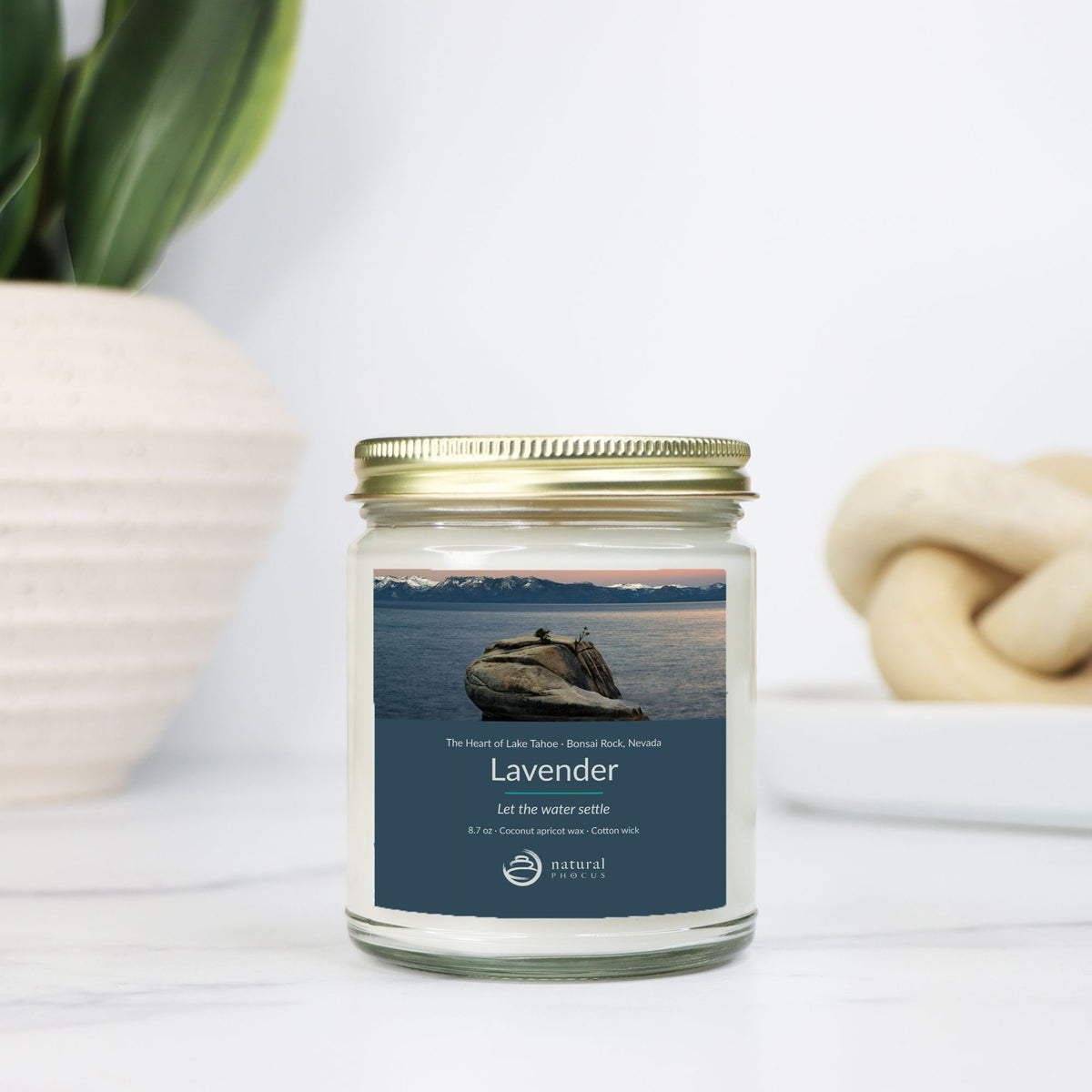 The Heart of Lake Tahoe Candle in Lavender, a clear glass jar candle with a gold lid, labeled with a photograph of Bonsai Rock in still water beneath snow-covered peaks at Lake Tahoe, Nevada