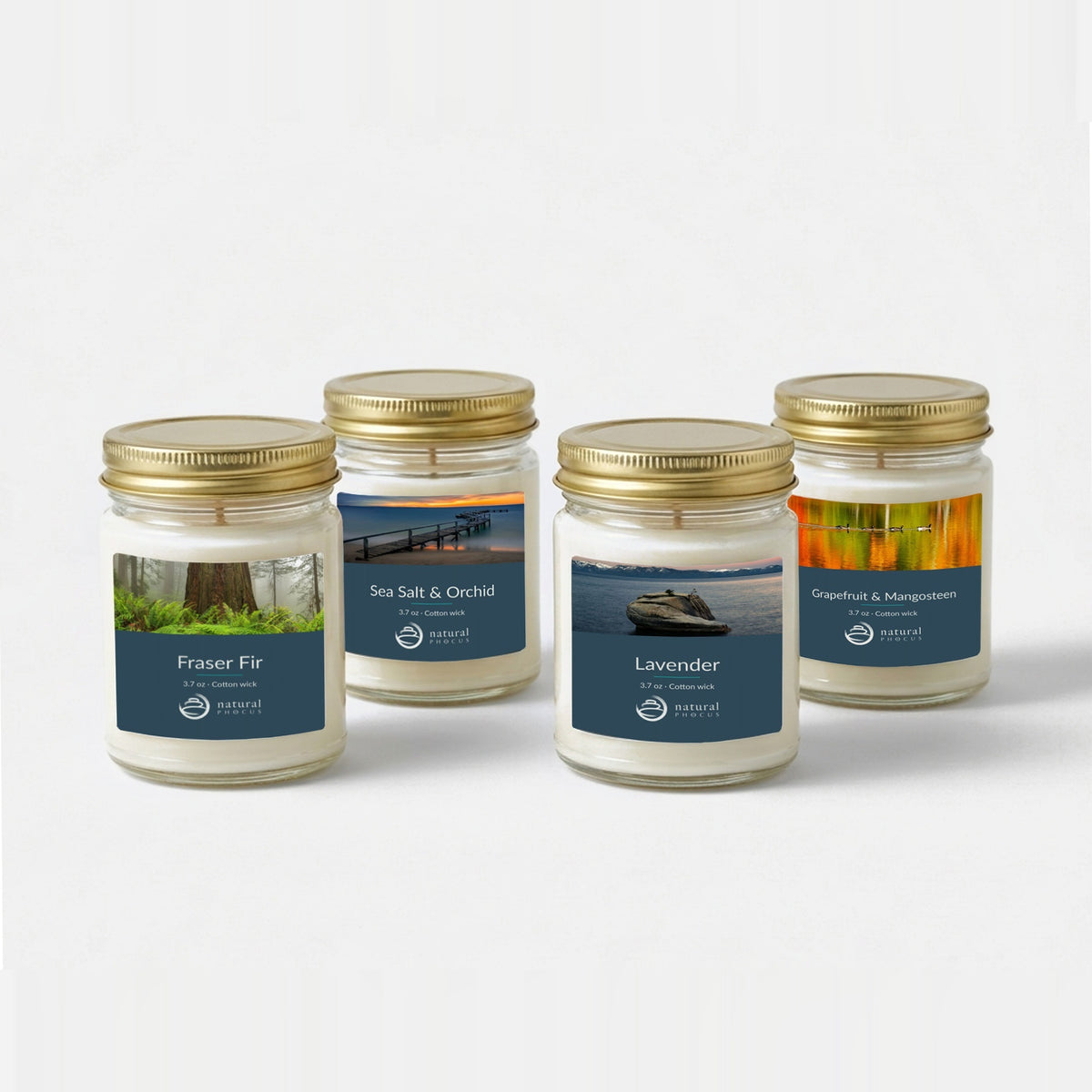 Everyday Discovery Set: four 4 oz clear glass mini candles with gold lids and navy labels, in Fraser Fir, Sea Salt and Orchid, Lavender, and Grapefruit and Mangosteen, each labeled with a landscape photograph