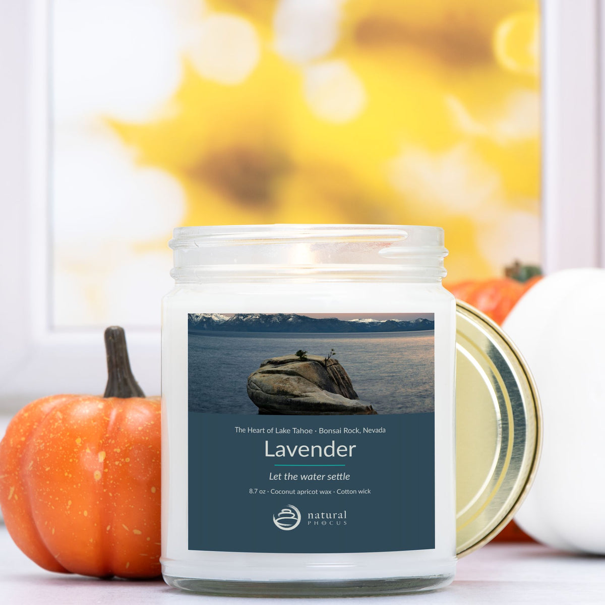 The Heart of Lake Tahoe Candle in Lavender, a clear glass jar candle with a gold lid, labeled with a photograph of Bonsai Rock in still water beneath snow-covered peaks at Lake Tahoe, Nevada