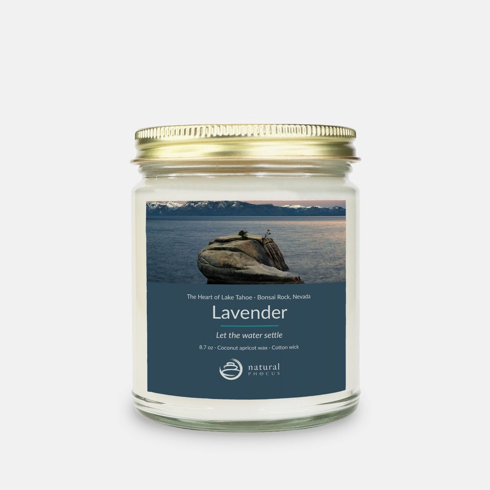 The Heart of Lake Tahoe Candle in Lavender, a clear glass jar candle with a gold lid, labeled with a photograph of Bonsai Rock in still water beneath snow-covered peaks at Lake Tahoe, Nevada