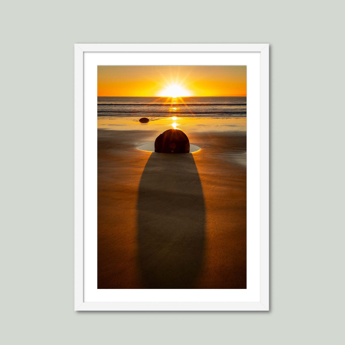 The Patient Stone, a Moeraki Boulder casting a long shadow toward the viewer as the sun rises over the Pacific in Otago, New Zealand, in a white frame with a white mat