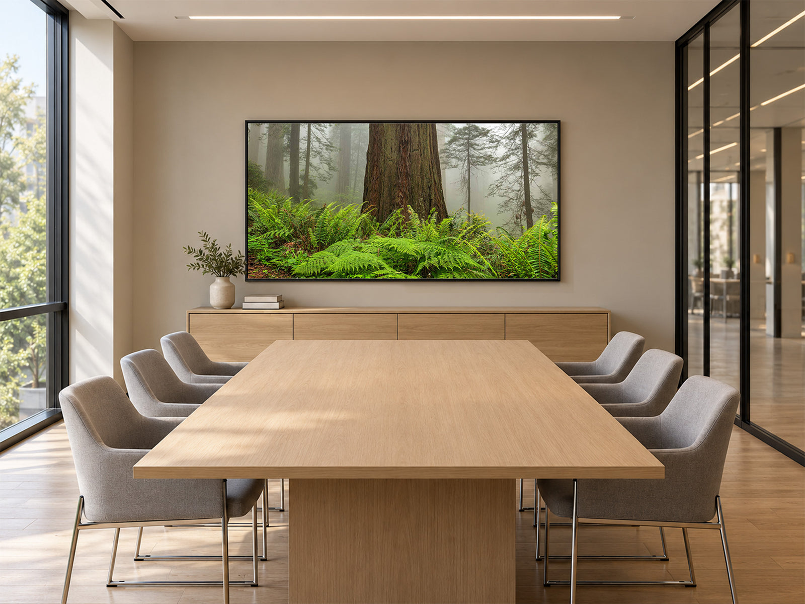 A modern boardroom with a light oak conference table and gray chairs facing Rooted, a photograph of a redwood rising from ferns in the mist, in a black frame